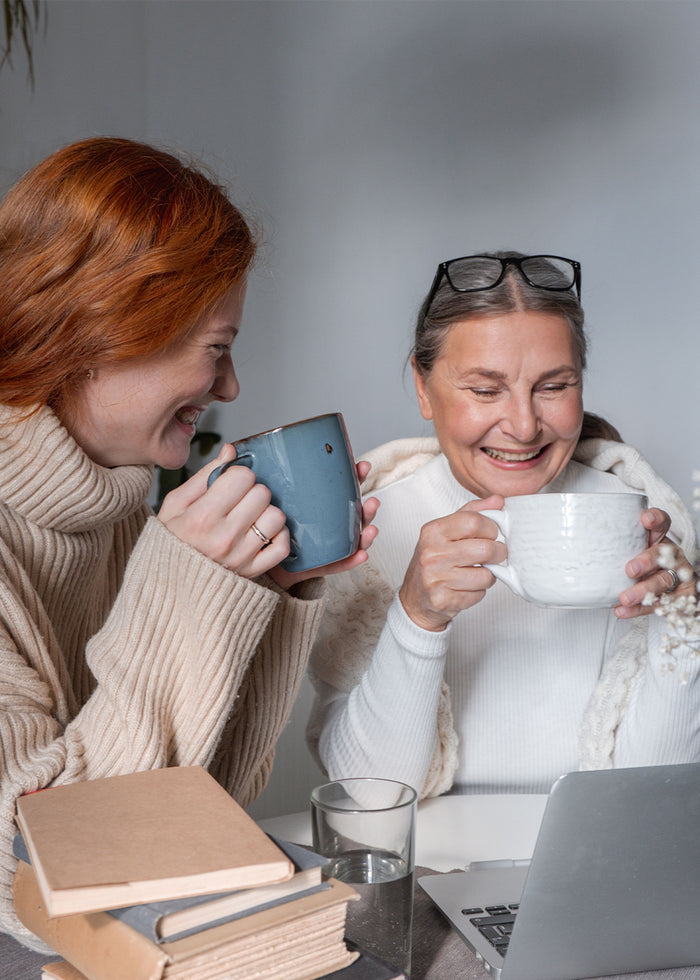 Zwei Frauen lachen gemeinsam, jede mit einer Tasse in der Hand.