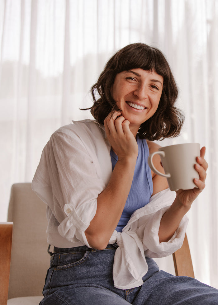 Eine Frau sitzt lächelnd mit einer Tasse in der Hand am Fenster.