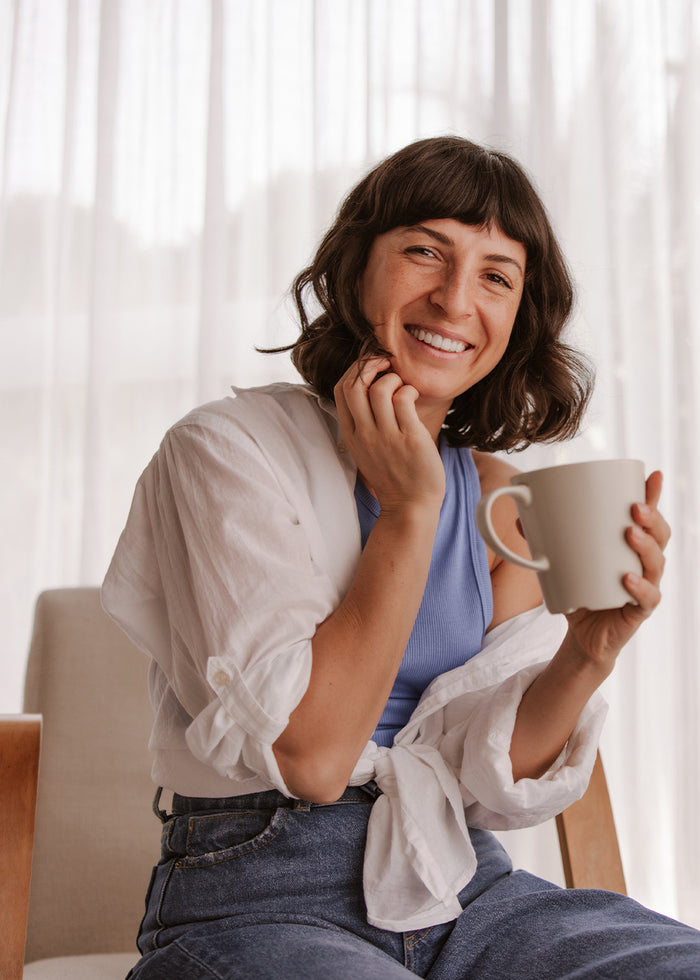Eine Frau sitzt lächelnd mit einer Tasse in der Hand am Fenster.
